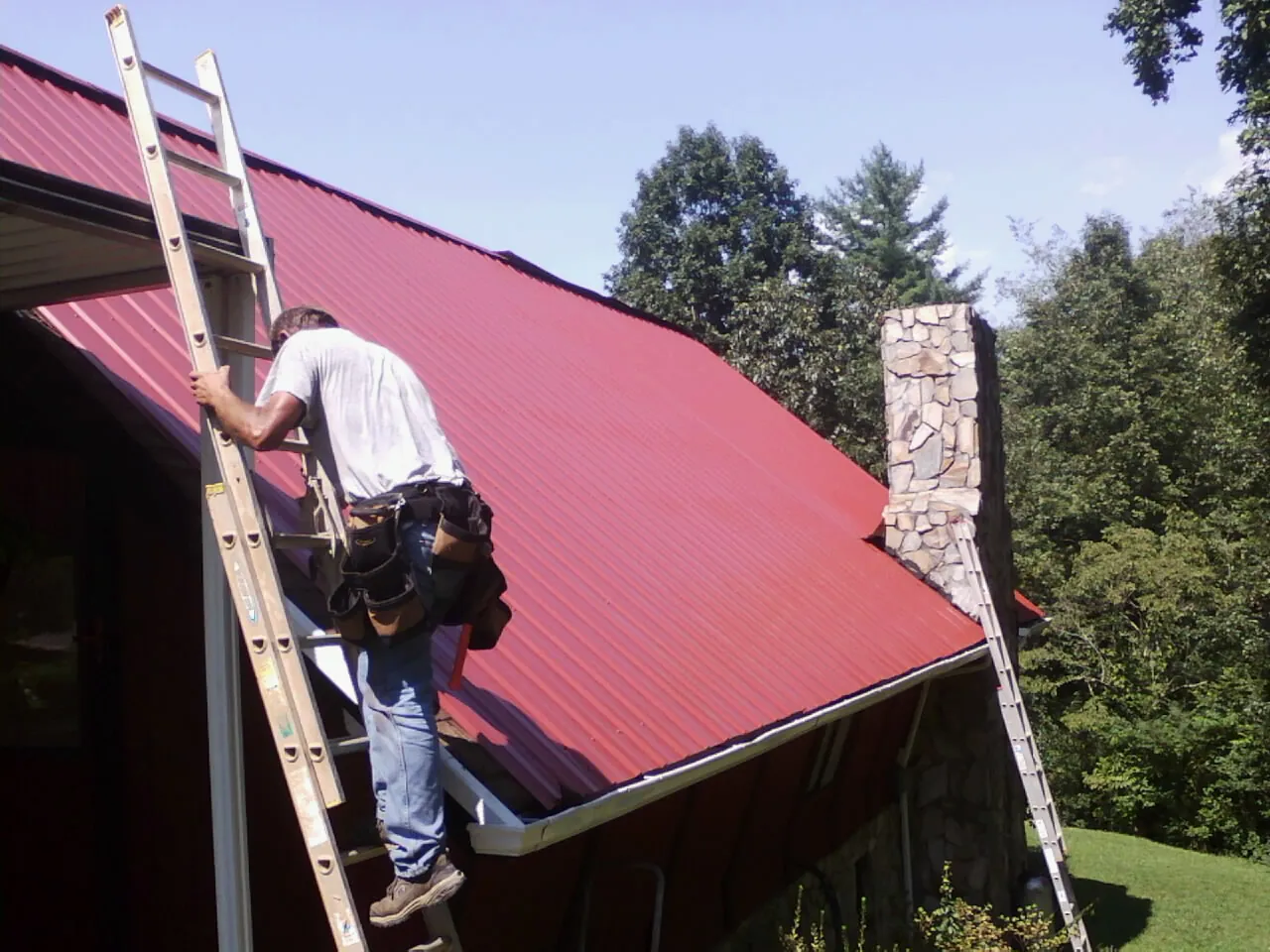 Roofer on a ladder beside a red metal roof and stone chimney — Soffit & Fascia Repair by Roofing Brothers in Poulan